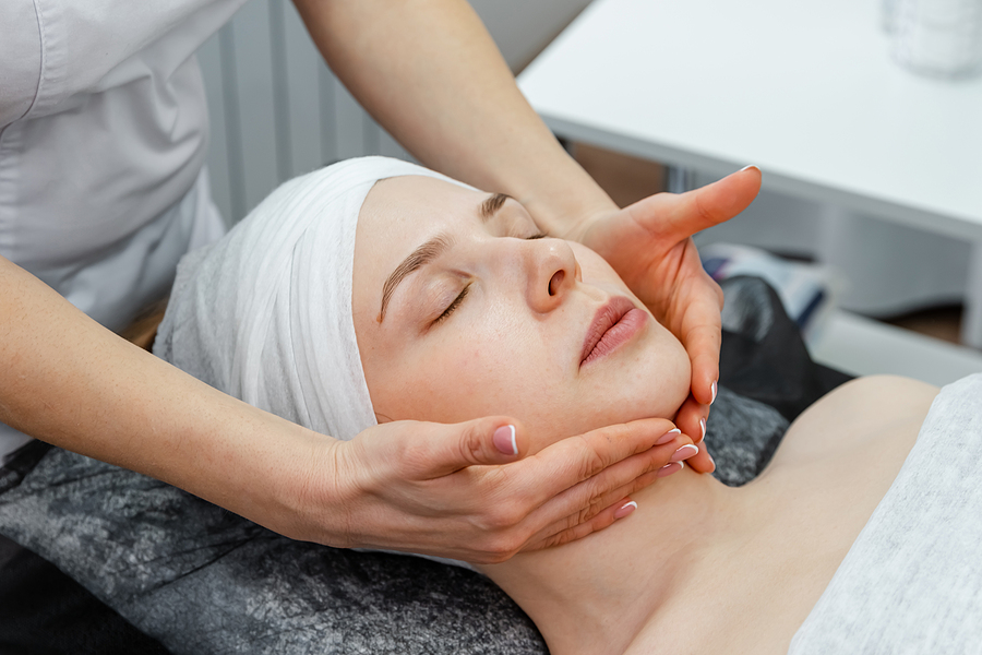 Professional beautician during the seance of facial massage. Charming young woman having face massage at beauty salon. Woman under facial treatment at beauty spa. Procedure of facial massage.